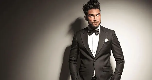A man in a black tuxedo and bow tie against a grey wall