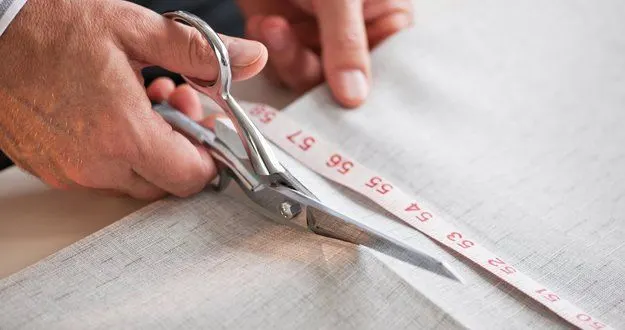A tailor's hands cutting pale fabric along a measuring tape